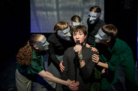 Die Theatergruppe Stellwerk Weimar mit ihrem Stück "Wilhelm Tell – Versuche". Zu sehen ist eine Person in der Mitte mit einem Mikro. Die Person wird von fünf anderen Schauspielern beträgt. Dabei tragen sie graue Masken.