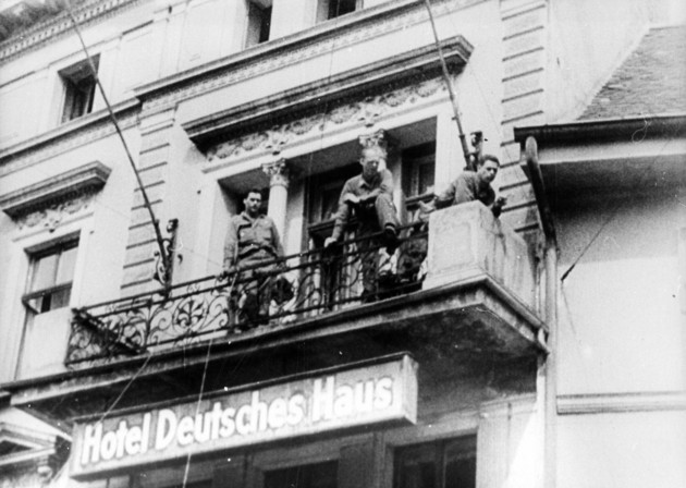 Drei Soldaten blicken nach unten von einem Balkon mit Schriftzug Hotel Deutsches Haus