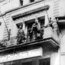 Drei Soldaten blicken nach unten von einem Balkon mit Schriftzug Hotel Deutsches Haus