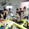 Jugendliche kochen gemeinsam, tragen bunte Schürzen, lesen an hellgrünen Arbeitsplatten Kochrezepte und scherzen