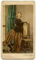 Frau mit üppigem Rock und dunkler Jacke steht seitlich an einen Stuhl gelehnt, ein großer schwarzer Hund liegt zu ihren Füßen