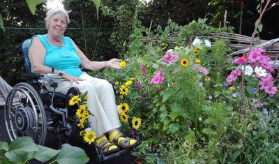 Foto von Christel Lehmann. Eine ältere Dame sitzt in einem Rollstuhl, lächelt in die Kamera und berührt eine gelbe Blume. Sie ist in einem Garten, welcher voller verschiedener Blumen ist.
