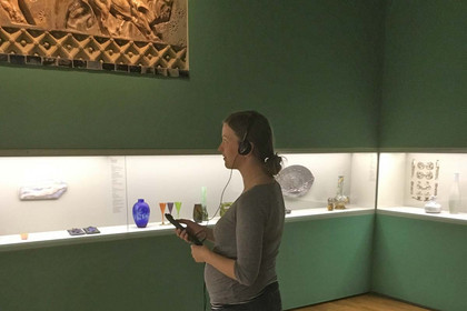 Eine Frau steht in einem Ausstellungsraum mit Kopfhörern und wählt über eine Fernbedienung den passenden Beitrag aus.