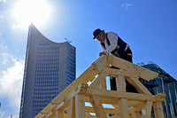 Ein Zimmermann werkelt auf einem Dachstuhl, im Hintergrund das City-Hochhaus