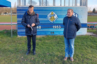 Heiko Rosenthal und Ralf Wittke stehen auf dem Rasen eines Sportplatzes vor einer Kabine, auf der das Vereinswappen zu sehen ist. 