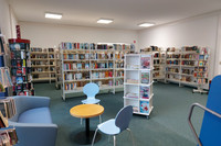 Im Eingangsbereich der Bibliothek steht links ein gemütlicher blauer Sessel an einem gelben runden Tisch, an dem noch zwei weitere Stühle stehen. Im Hintergrund stehen Regale mit Büchern. 