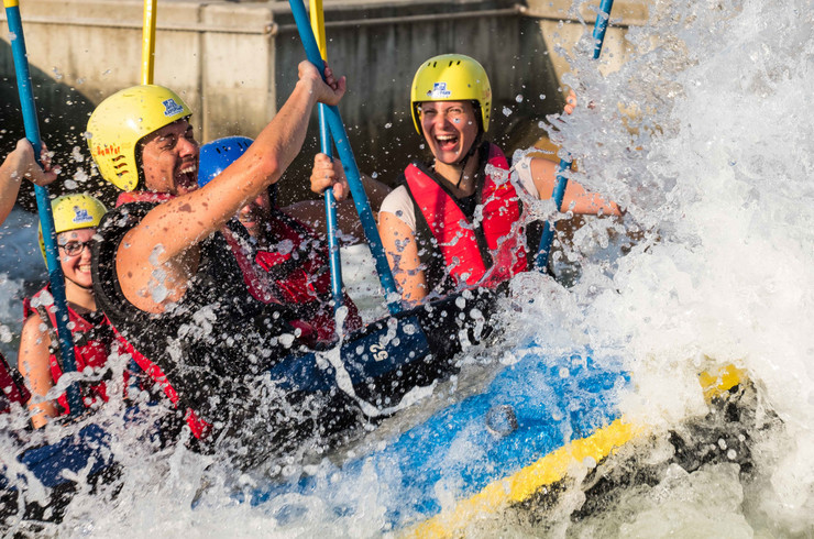 Menschen mit Helm und Paddel fahren lachend in einem Raftingboot durch wildes Gewässer.