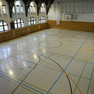 Sporthalle mit verschiedenen Bodenmarkierungen für unterschiedliche Sportarten und einem Basketballkorb an einer Wand.