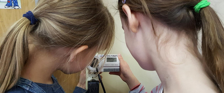 Zwei Mädchen - von hinten fotografiert - schauen auf ein Stromverbrauchsmessgerät, um den gemessenen Wert abzulesen.