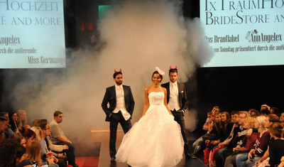 Hochzeitsmode wird von einer Braut und zwei Männern im Anzug auf einer Modenschau präsentiert.