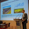 Martin Faßhauer von den Stadtwerken Leipzig spricht zum Thema erneuerbare Energieerzeugung in Leipzig. Dabei steht er am Rednerpult auf der Bühne und im Hintergrund ist seine Präsentation zu sehen.