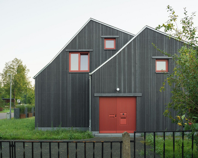 Ansicht einer mit Holz verkleideten Hausfassade mit Doppelgiebel, einer roten Eingangstür und roten Fensterrahmen.