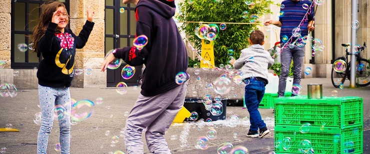 Viele Seifenblasen schweben über der Straße. Drei Kinder versuchen diese zu fangen. Ein Erwachsener ist zu sehen, der im Hintergrund die Seifenblasen entstehen lässt.