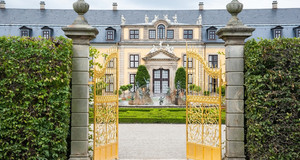 Ein geöffnetes goldenes Tor gibt den Blick frei auf einen Springbrunnen im Vordergrund und ein gelbes Schloss mit schwarzem Dach im Hintergrund.