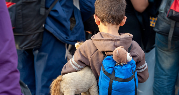 Ein Kind steht hinter eine Gruppe von Menschen mit Gepäck auf dem Rücken und einem Plüschtier unter dem Arm.