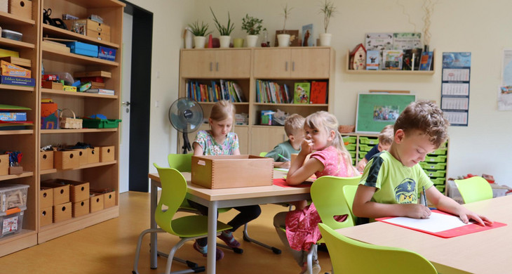An zwei Tischen sitzen mehrere Jungen und Mädchen und malen. an den Wänden sind Regale mit Spielzeugen, Büchern und Bastelmaterial aufgestellt.