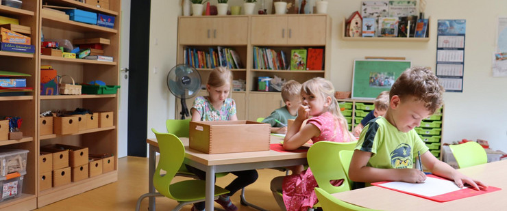 An zwei Tischen sitzen mehrere Jungen und Mädchen und malen. an den Wänden sind Regale mit Spielzeugen, Büchern und Bastelmaterial aufgestellt.