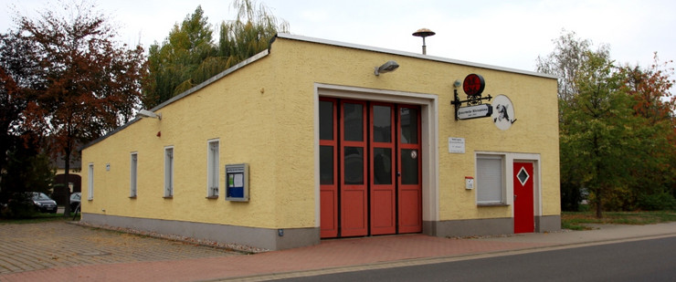 Eine gelb angestrichene Garage mit einem großen, roten Tor. Die Garage wird nach hinten hin flacher. Neben dem Tor sind ein Fenster und eine rote Eingangstür.