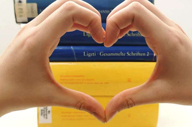 Zwei Hände formen ein Herz im Vordergrund, im Hintergrund ein Stapel Bücher in den ukrainischen Landesfarben Blau und Gelb