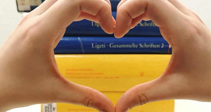 Zwei Hände formen ein Herz im Vordergrund, im Hintergrund ein Stapel Bücher in den ukrainischen Landesfarben Blau und Gelb