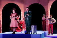 Mehrere Schauspieler auf der Bühne eines Theaters. Alle tragen rote und rosa Sachsen, bis auf einen Mann mit dunklen Anzug und einem Reisekoffer.