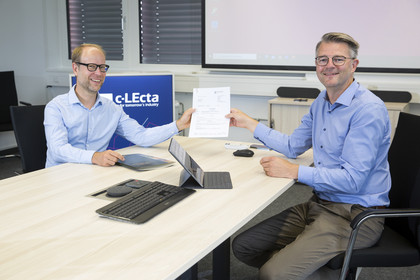 Amtsleiter der Wirtschaftsförderung, Clemens Schülke in hellblauem Hemd übergibt Geschäftsführer von C-LEcta, Dr. Marc Struhalla, den Fördermittelbescheid. Beide sitzen an einem Tisch auf dem ein Laptop und eine Tastatur liegen.
