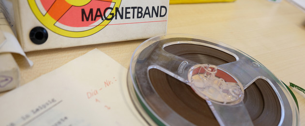 Ein Tonband auf einem Tisch, dahinter ein Karton mit der Aufschrift "Magnetband"