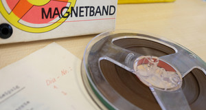 Ein Tonband auf einem Tisch, dahinter ein Karton mit der Aufschrift "Magnetband"