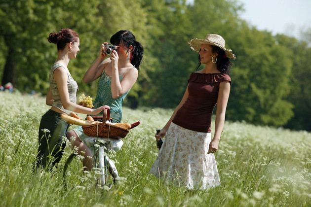 Drei Frauen auf einer Wiese. Eine der Frauen fotografiert eine Frau mit Fahrrad