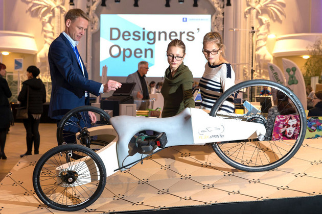 Ausstellungsstück auf der Designers Open: Ein futuristisch anmutendes Dreirad. Dahinter stehen Menschen und schauen es sich an.