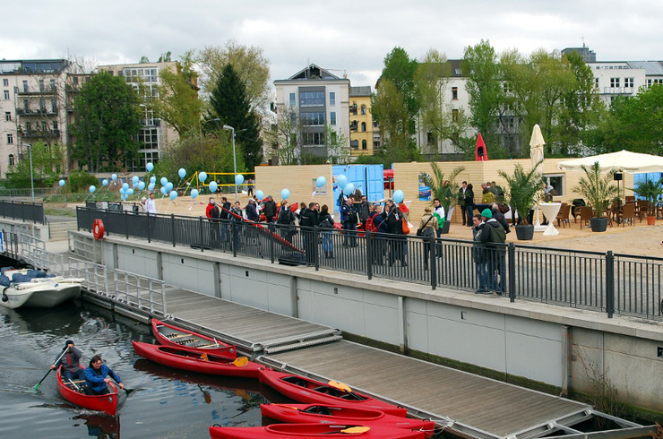 Fünf rote Kajaks, teils mit Menschen besetzt, befinden sich im Wasser. Etwa einen Meter erhöht befindet sich der Uferbereich mit Geländer und Schaulustigen.