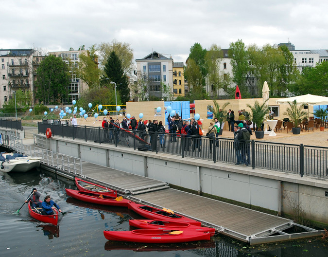 Fünf rote Kajaks, teils mit Menschen besetzt, befinden sich im Wasser. Etwa einen Meter erhöht befindet sich der Uferbereich mit Geländer und Schaulustigen.