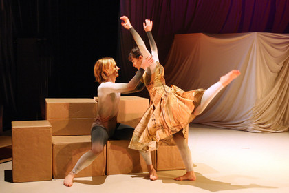 Ein Tänzer sitzt auf Pappkartons und hält eine Tänzerin die vor ihm steht.