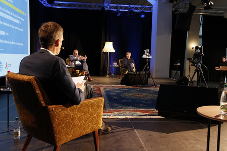 Blick auf ein Podium, auf dem drei Männer beim diskutieren zu sehen sind.