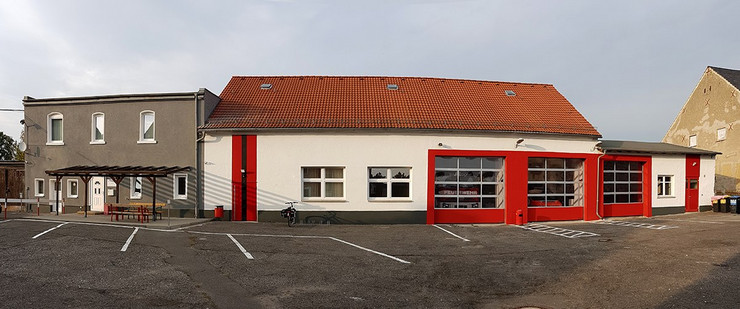 Eine eingeschossige Fahrzeughalle mit drei großen Ausfahrten, die mit roter Farbe umrandet sind. Links neben der Halle zwei Fenster und eine rote Eingangstür. Daran angebaut ist ist ein zweigeschossiges, graues Haus.
