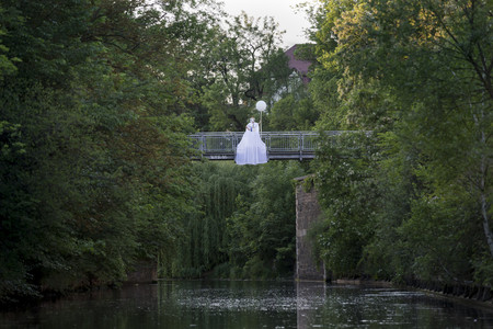 Ein Mensch in langem weißem Kleid mit Maske und weißem Ballon in der Hand sitzt auf einem Brückengeländer.