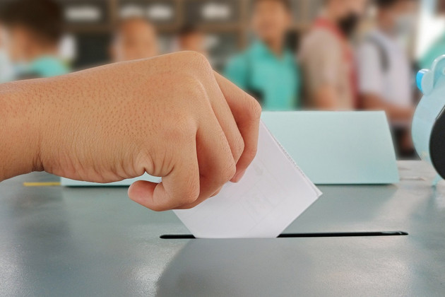 Eine Kinderhand mit einem Stimmzettel wirft diesen in eine Wahlurne.
