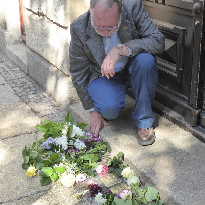 Ein älterer Herr mit Kippa hockt vor einem Hauseingang und legt Blumen zu zwei goldenen Stolpersteinen.