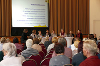 Mehrere Leute sitzen vor Publikum in einer Diskussionsrunde. Auf einer Leinwand im Hintergrund sind die anwesenden Teilnehmer aufgelistet.