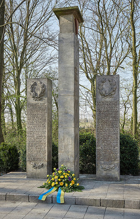 Vor dem Denkmal der 53 liegt ein Blumenkranz. Das Denkmal besteht aus drei aufragenden Säulen.