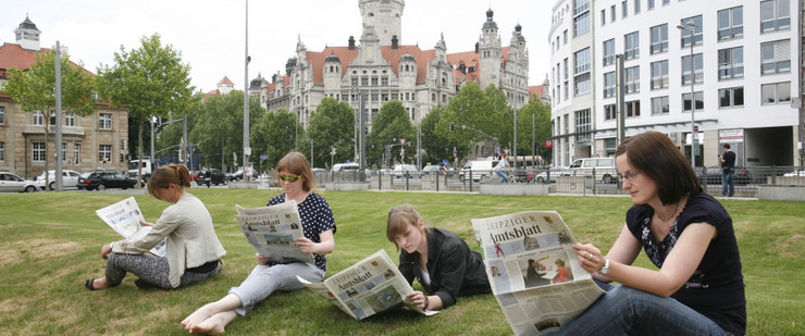 Vier Menschen lesen auf einer Wiese Amtsblatt. Im Hintergrund ist das Neue Rathaus.
