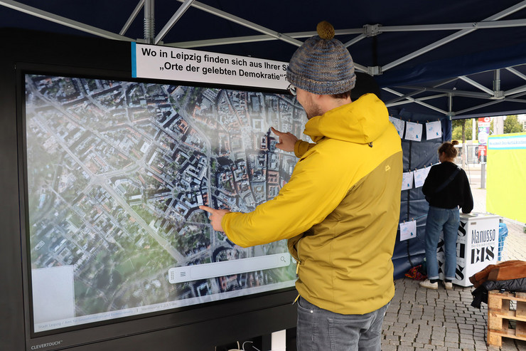 Ein Mann in gelber Jacke steht vor einem großen Bildschirm, auf dem ein Stadtplan von Leipzig abgebildet ist
