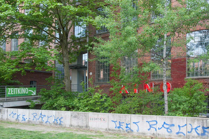 Eine niedrige Betonmauer mit Graffiti besprüht, im Hintergrund ein Backsteinhaus mit großem Schriftzug "Kino", halb von Bäumen verdeckt