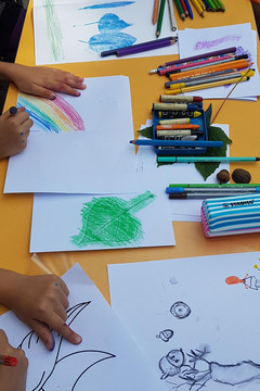 Auf dem Bild sind Kinderhände zu sehen, die auf einem Tisch Bilder ausmalen oder selbst malen, beispielsweise einen Regenbogen. Auf dem Tisch liegen außerdem verschiedenste Stifte (Filzstifte, Wachsmalstifte, Buntstifte und so weiter).