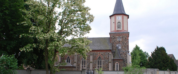 Eine Kirche umgeben von einer Mauer. Davor ein blühender Robinienbaum und eine kleine Fußgängerbrücke aus Metall.