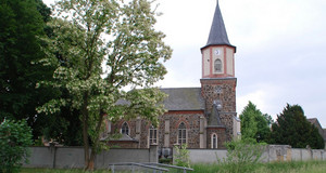 Eine Kirche umgeben von einer Mauer. Davor ein blühender Robinienbaum und eine kleine Fußgängerbrücke aus Metall.