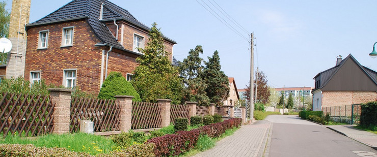 Eine Straße in Miltitz mit dörflichen Einfamilienhäusern aus Backstein. Die Plattenbauten Grünaus sind von Weitem zu sehen.