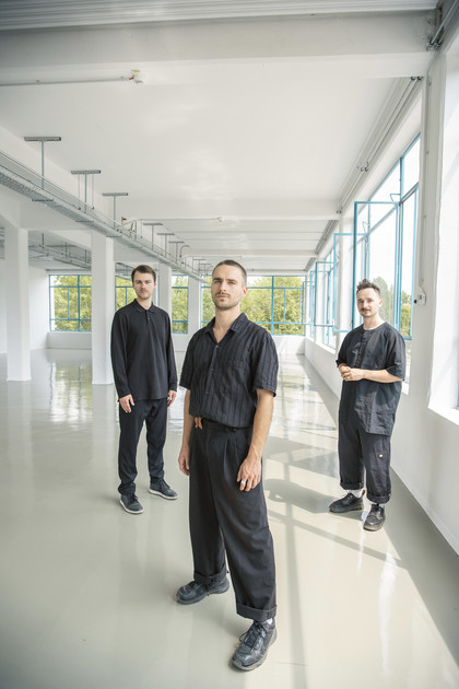Die drei Personen der Band Simon Lucacio Trio stehen schwarzgekleidet in einem lichtdurchfluteten weißen Raum.