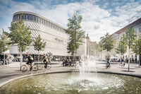 Richard-Wagner-Platz mit einem Springbrunnen im Vordergrund, mehreren Radfahrern und dem Einkaufszentrum Höfe am Brühl