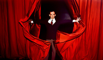 Foto vom Choreograf und Ballettdirektor Uwe Scholz. Ein Mann im Anzug blickt in die Kamera während er zwischen roten Vorhängen steht und diese zur Seite drückt.
